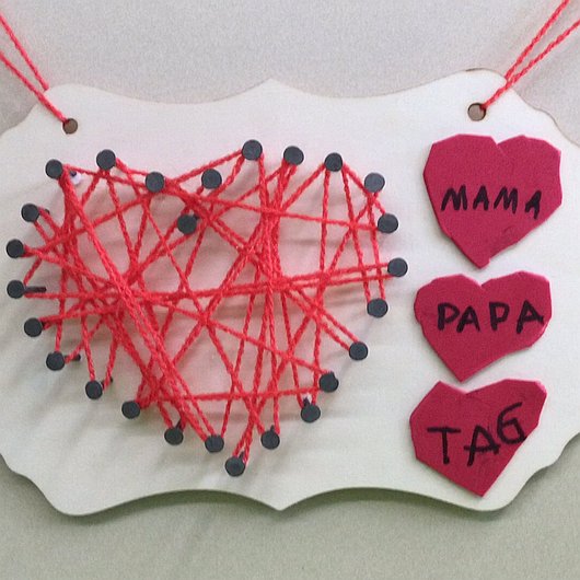 Herzförmiges Nagelbild mit roten Wollfäden und Schildern mit kleinen roten Schildchen mit den Texten „Mama“, „Papa“ und „Tag“