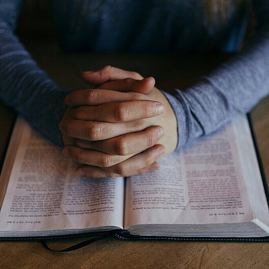 Gefaltete Hände auf einer offenen Bibel (Foto von Patrick Fore auf Unsplash)